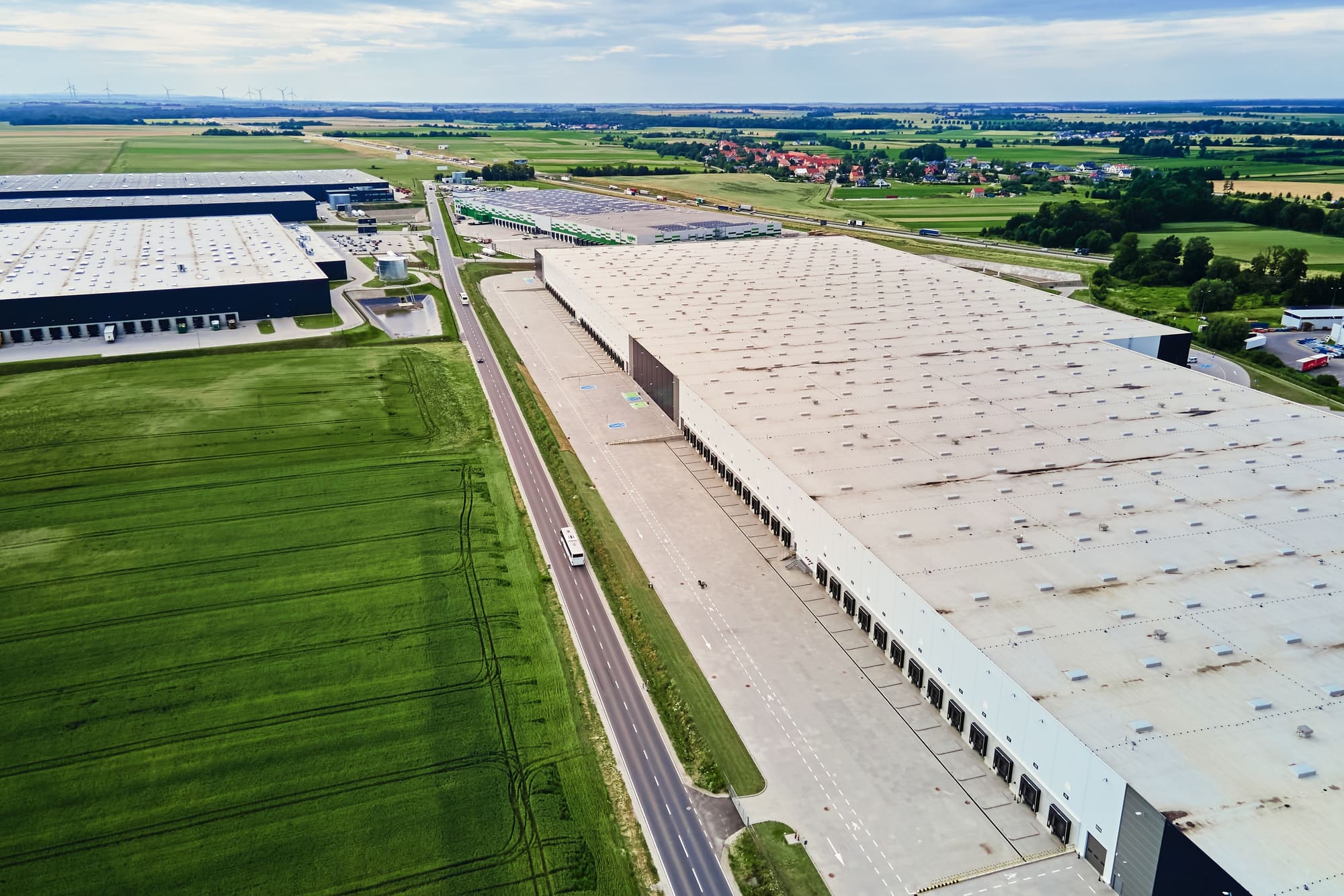 Luftbild einer großen Halle zwischen Feldern, dahinter ein Dorf